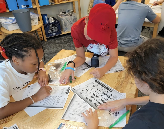 Students gathered around a classroom table comparing DNA gel patterns on printed worksheets